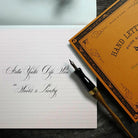 A close-up styled flat-lay showing a black and gold fountain pen resting on white lined paper with elegant cursive writing, next to an orange book titled 'Hand Lettering Guide'.