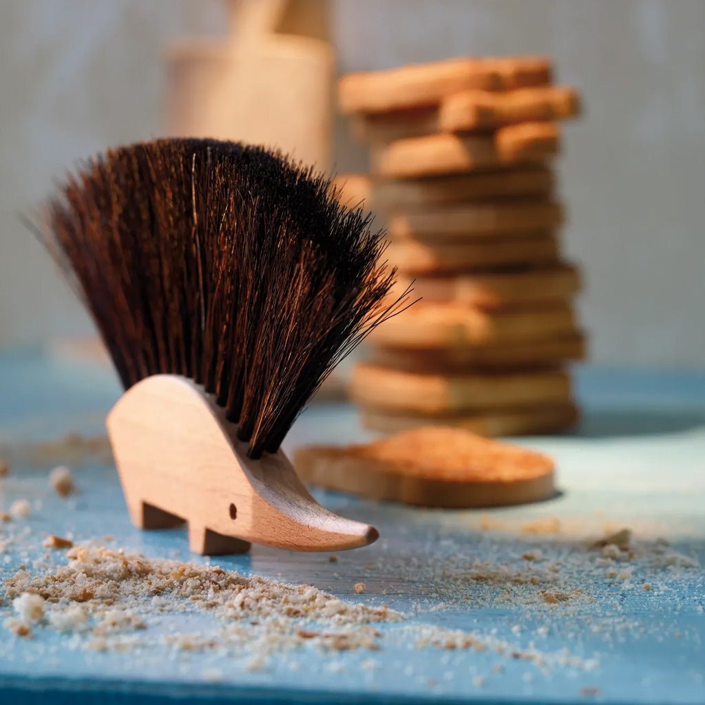 A small wooden hedgehog-shaped brush with dark brown bristles stands on a light blue surface with wood shavings and blurred wooden blocks in the background.