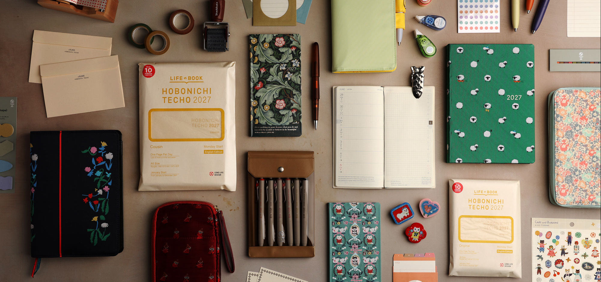 Overhead view of colorful Hobonichi 2027 planners, covers, pens, stickers, and stationery arranged on a tabletop.