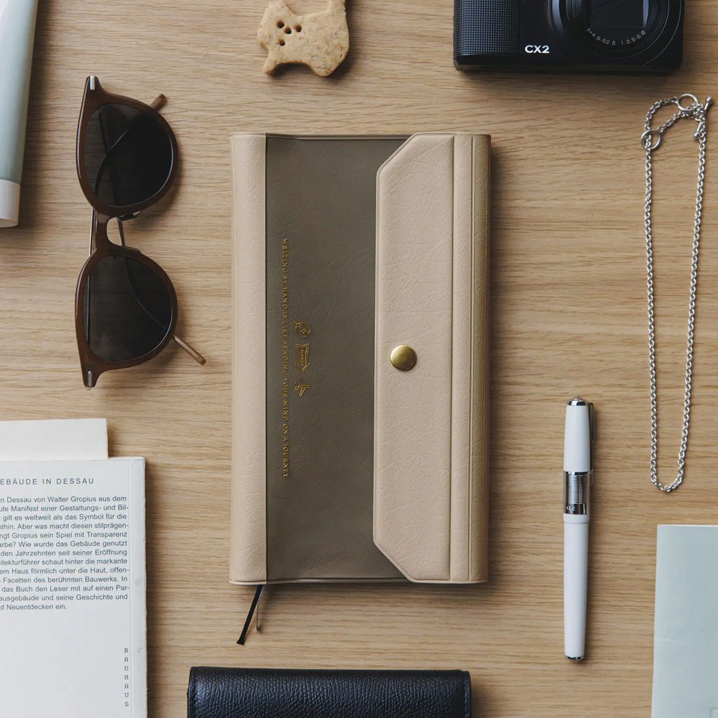 Light warm gray alettone case with front pocket and button closure for Hobonichi Techo Weeks on wooden desk with stationery accessories