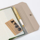 A beige folio-style notebook cover with a snap closure, shown open on a white background. Inside is a white dotted notebook, a gold-and-white pen, and a small brown card with black printed graphics.