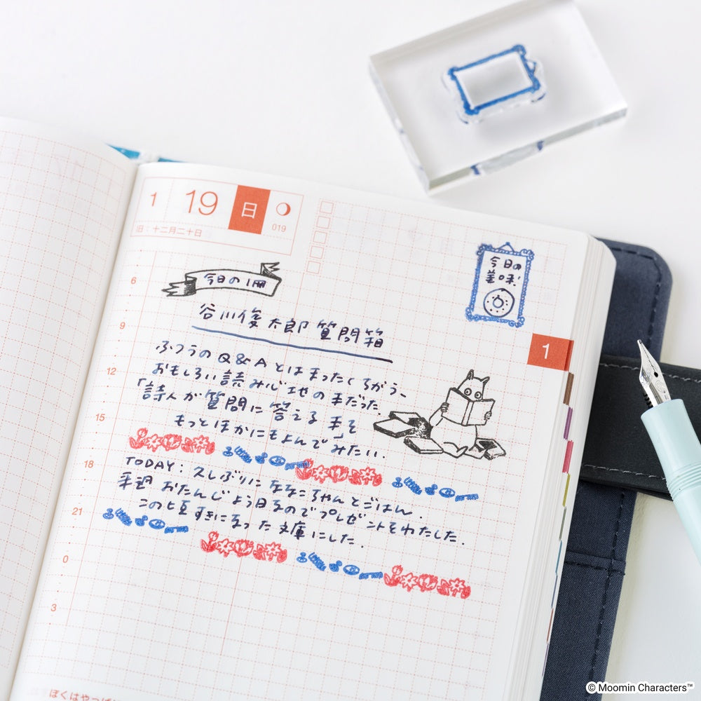 An open planner or journal page with handwritten Japanese notes, a small Moomin stamp design, and colored date markers, styled on a white surface with a blue pen, a dark folio, and a clear stamp box.