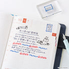 An open planner or journal page with handwritten Japanese notes, a small Moomin stamp design, and colored date markers, styled on a white surface with a blue pen, a dark folio, and a clear stamp box.