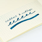 A close-up of a hand-written note on graph paper, showing the cursive text 'collect & collage' written in dark blue ink with a matching underline.