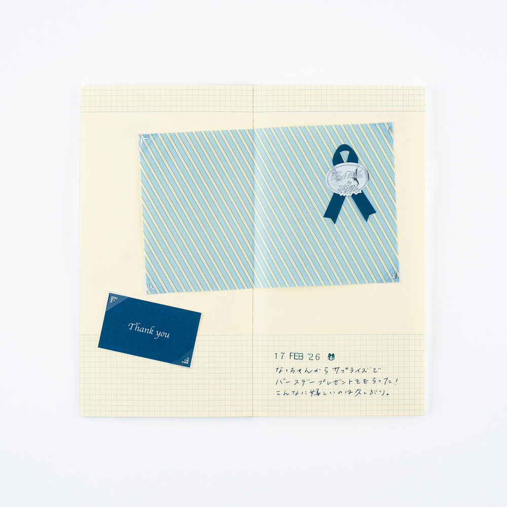 A gridded pasting notebook with a cream grid background, featuring a blue striped square, a blue ribbon emblem, and a small blue card reading 'Thank you'.
