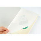 A hand opening a cream-colored notebook or journal, showing a pale yellow interior page and faint printed illustration.