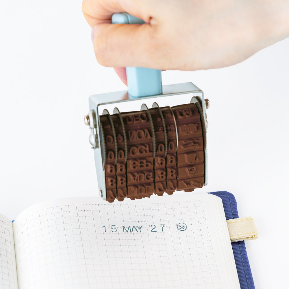 A hand holds a metal rotating stamp over an open grid notebook, showing the engraved date stamping action. The stamp has a light blue handle and a silver metal frame with dark brown engraved letter and number blocks.
