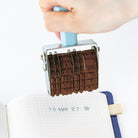 A hand holds a metal rotating stamp over an open grid notebook, showing the engraved date stamping action. The stamp has a light blue handle and a silver metal frame with dark brown engraved letter and number blocks.