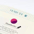 A close-up of a cream-colored gridded page featuring a pink and purple circular logo, the word 'Handwrite', and a black ink nib illustration. The top of the light blue graph paper shows the date '18 NOV '26'.