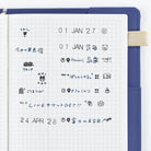 A blue notebook cover with a white grid page insert, showing handwritten Japanese notes and small stamped icons in blue ink. 