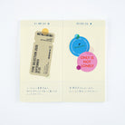 A two-page planner spread or notebook spread on a white background. The pages are cream-colored with a light yellow grid pattern, featuring a paper ticket graphic, blue and pink circular stickers, and handwritten Japanese text.