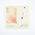 A pocket notebook with a cream-colored grid page, featuring a pastel peach watercolor illustration on the left, handwritten notes in Japanese, and a white bookmark with black calligraphy.
