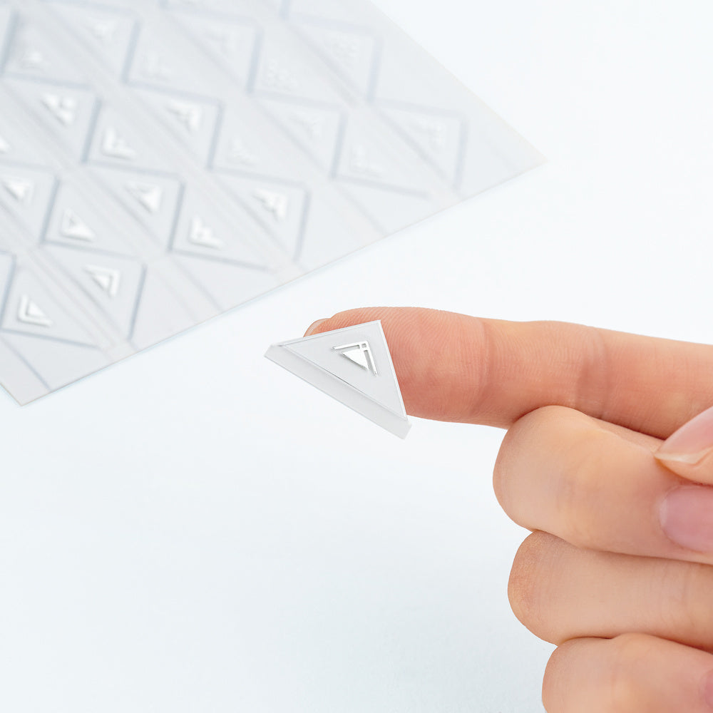 A close-up of a hand holding a small triangular white sticker, with a sheet of similar triangular pieces blurred in the background.