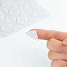 A close-up of a hand holding a small triangular white sticker, with a sheet of similar triangular pieces blurred in the background.