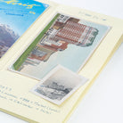 A vintage-style travel postcard album page with handwritten notes, featuring a large tilted postcard of a red-roofed building and a smaller black-and-white postcard of ships, alongside a partial blue postcard in the corner.