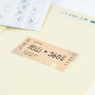 A close-up of a station ticket style sticker or label on a pale yellow background, with part of a sticker sheet and a lined paper visible behind it.