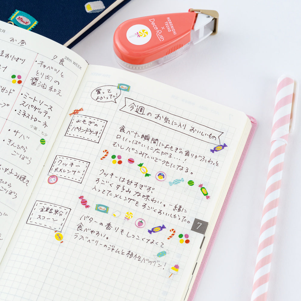An open notebook with handwritten Japanese notes and colorful candy tape designs, styled on a white surface with a pink-and-white striped pen, a coral tape dispenser, and part of a blue notebook visible.