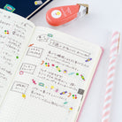 An open notebook with handwritten Japanese notes and colorful candy tape designs, styled on a white surface with a pink-and-white striped pen, a coral tape dispenser, and part of a blue notebook visible.