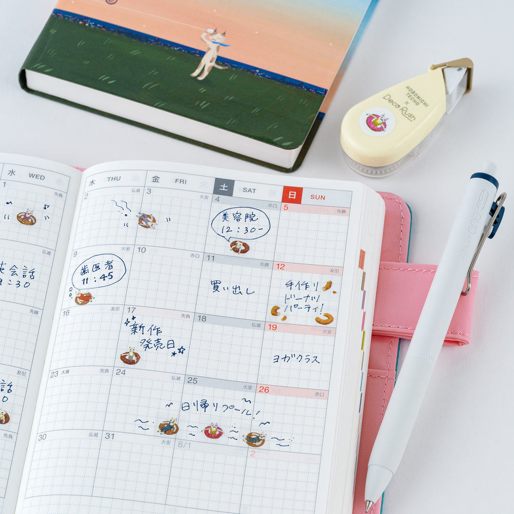 An open weekly planner with Japanese handwriting and small cat and donut illustrations, styled on a white surface with a pink pen loop, white pen, cream correction tape, and a green notebook in the background.