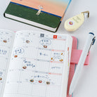 An open weekly planner with Japanese handwriting and small cat and donut illustrations, styled on a white surface with a pink pen loop, white pen, cream correction tape, and a green notebook in the background.