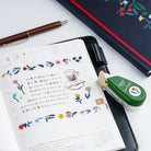 An open planner or notebook with Japanese writing and colorful floral deco rush tape, styled on a white surface with a pen, a green tape dispenser, and a dark patterned notebook.