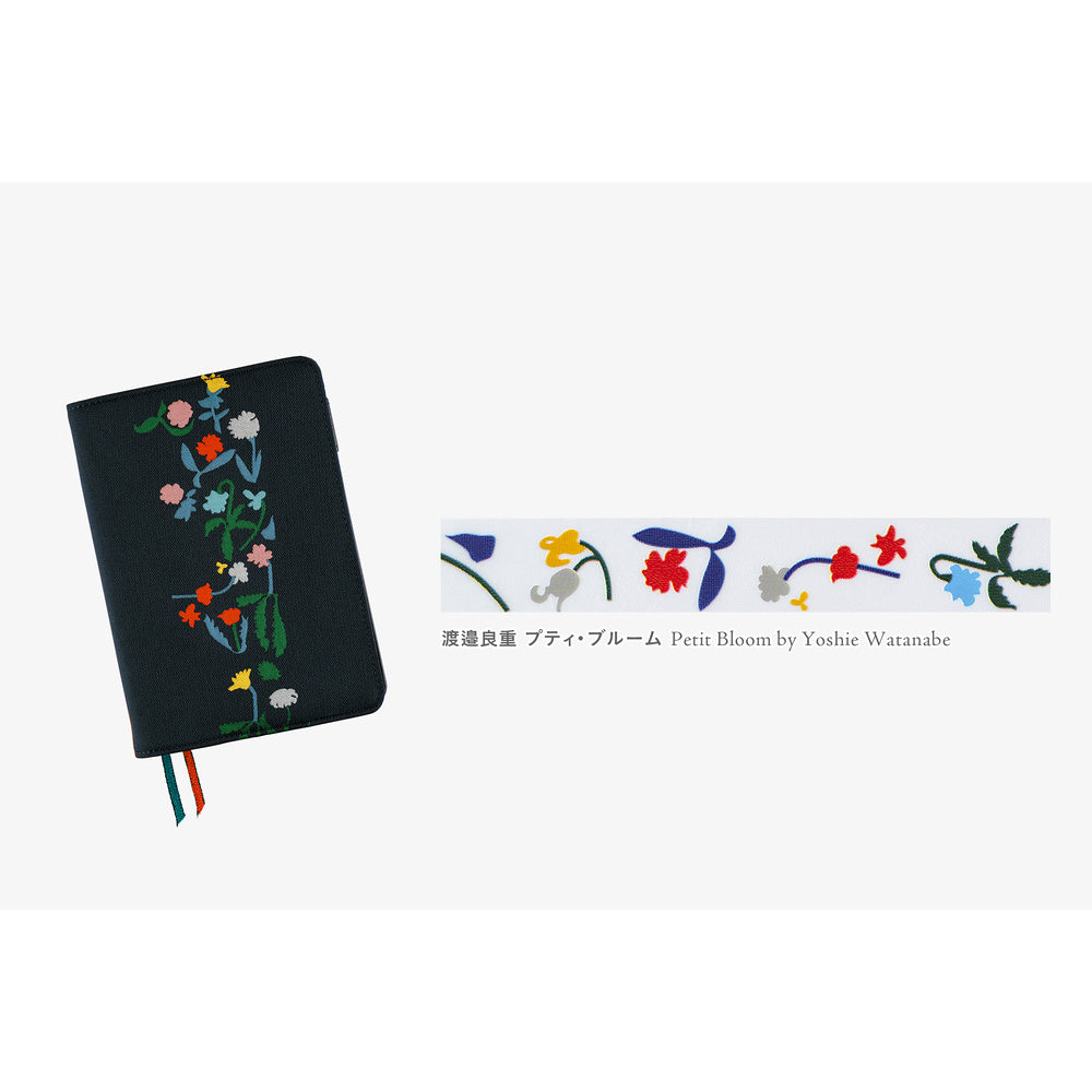 A black notebook cover with colorful floral embroidery on the front, shown on a light gray background. To the right is a close-up strip of floral deco rush tape with Japanese text and 'Petit Bloom by Yoshie Watanabe'.