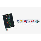 A black notebook cover with colorful floral embroidery on the front, shown on a light gray background. To the right is a close-up strip of floral deco rush tape with Japanese text and 'Petit Bloom by Yoshie Watanabe'.