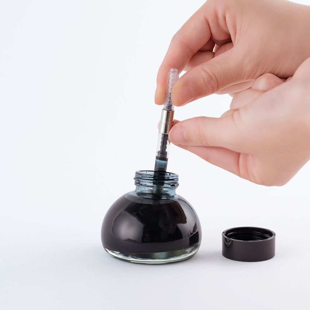 A hand is shown dipping a clear ink converter into a dark ink bottle with its cap removed, set against a clean white background.