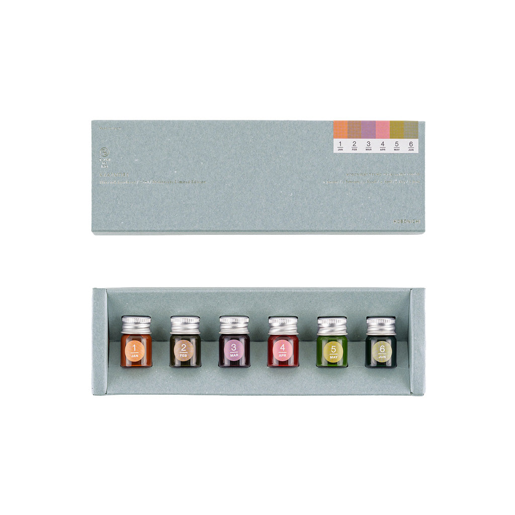A set of six small glass ink bottles with silver caps arranged in a light gray presentation box, shown below the matching box lid. The bottles have color labels numbered 1 through 6.