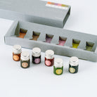 Six small glass bottles with silver caps, each containing a different colored ink, arranged in front of an open gray box with matching slots. The bottles are labeled 1 through 6 with circular stickers.
