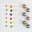 Six small glass bottles of ink with silver caps are arranged next to a white monthly planner page showing color swatches and handwritten notes in matching shades.
