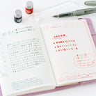 An open pink planner showing handwritten and printed Japanese notes, photographed on a white surface with a pen, two small bottles, and a glass quill nearby.