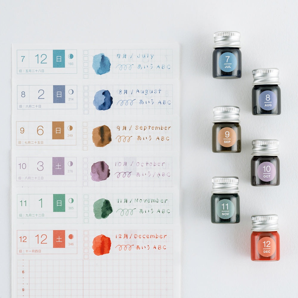 A flat-lay of a Japanese monthly calendar page next to twelve small glass ink bottles with silver caps, arranged on a clean white background. Each bottle is labeled with a number from 7 to 12 and a corresponding month name.