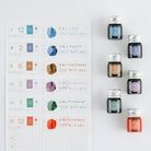 A flat-lay of a Japanese monthly calendar page next to twelve small glass ink bottles with silver caps, arranged on a clean white background. Each bottle is labeled with a number from 7 to 12 and a corresponding month name.