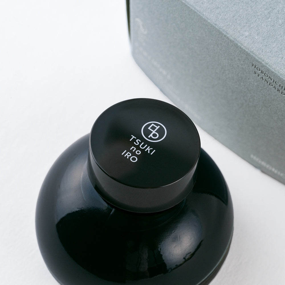 A close-up of a glossy dark blue bottle of ink with a matching black cap printed with the TSUKI no IRO logo, set against a light gray background with part of a gray box visible.