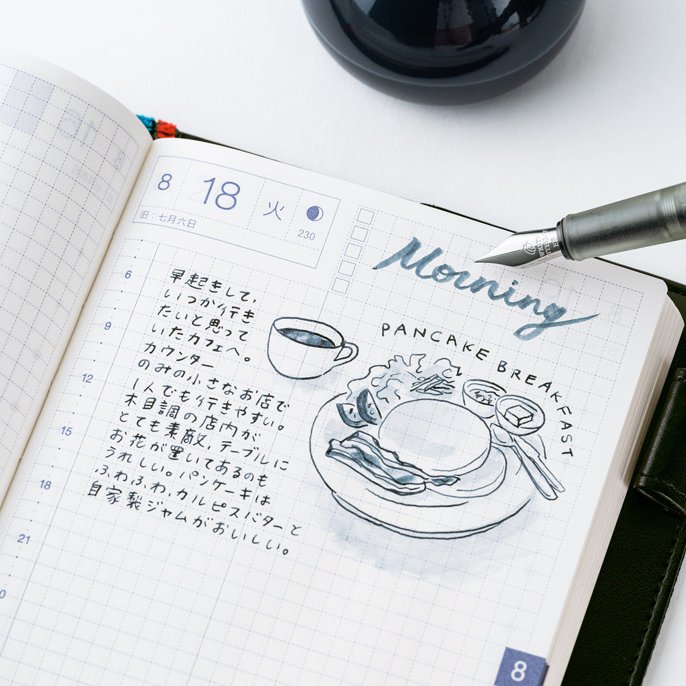 An open grid planner or journal showing a decorated morning log with handwritten text, a coffee cup sketch, and the words 'Morning Pancake Breakfast.' A silver pen rests on the page, and part of a dark round object is visible at the top against a white background.