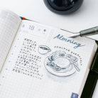 An open grid planner or journal showing a decorated morning log with handwritten text, a coffee cup sketch, and the words 'Morning Pancake Breakfast.' A silver pen rests on the page, and part of a dark round object is visible at the top against a white background.