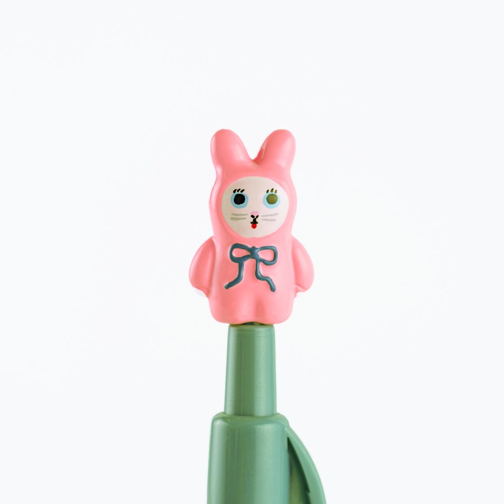 A close-up of a pink bunny-shaped pen topper with a blue bow and painted face, mounted on a green cylindrical body against a plain white background.