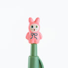 A close-up of a pink bunny-shaped pen topper with a blue bow and painted face, mounted on a green cylindrical body against a plain white background.
