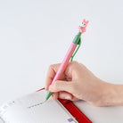 A hand holding a pink retractable pen with a green clip and a small pink cartoon character on top, writing in a lined notebook with a red pen loop visible.