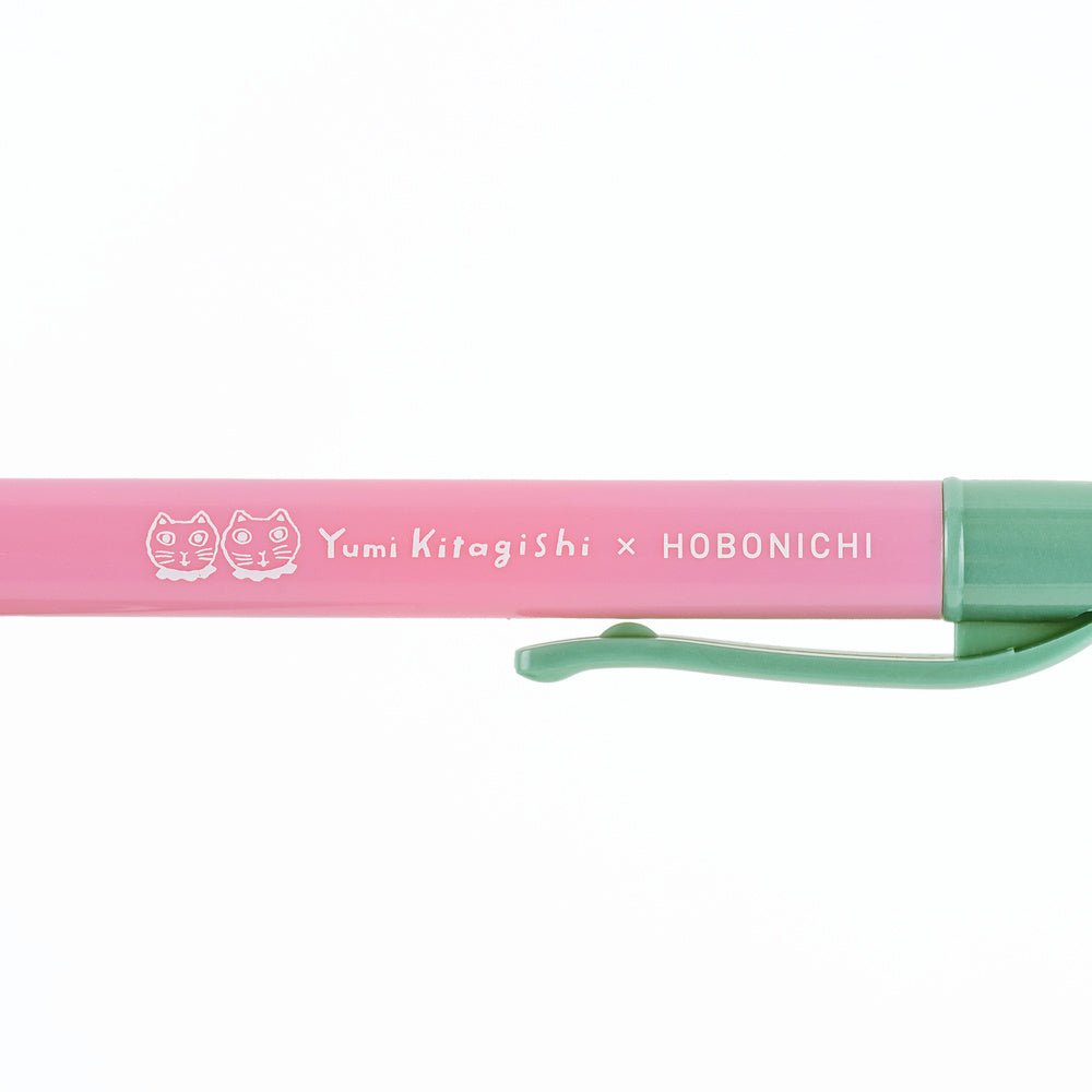 A close-up of a pink and mint green pen with white printed text reading 'Yumi Kitagishi x HOBONICHI' and two cat face icons, shown on a plain white background.