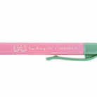 A close-up of a pink and mint green pen with white printed text reading 'Yumi Kitagishi x HOBONICHI' and two cat face icons, shown on a plain white background.