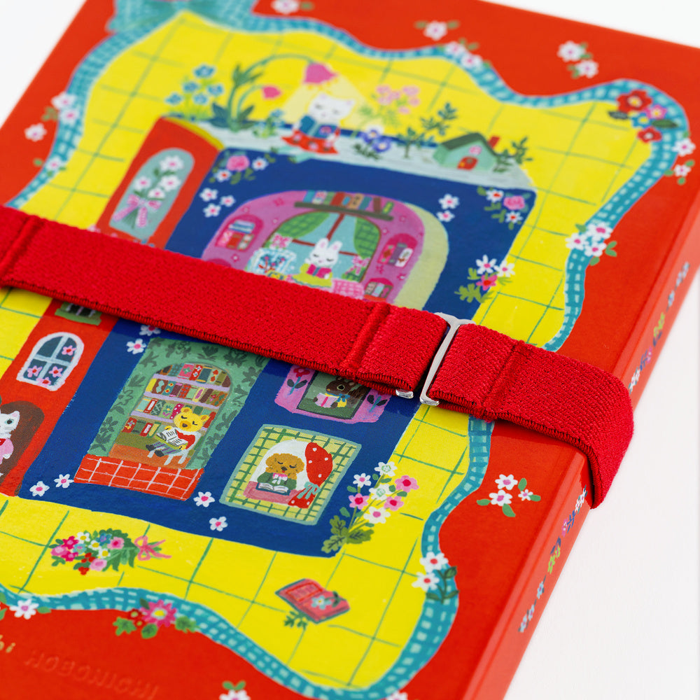 A close-up of a colorful illustrated book or notebook cover wrapped with a red elastic book band. The cover features a bright yellow grid background with a whimsical house illustration in pink, blue, green, and red, plus small white flowers.