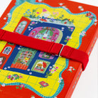 A close-up of a colorful illustrated book or notebook cover wrapped with a red elastic book band. The cover features a bright yellow grid background with a whimsical house illustration in pink, blue, green, and red, plus small white flowers.