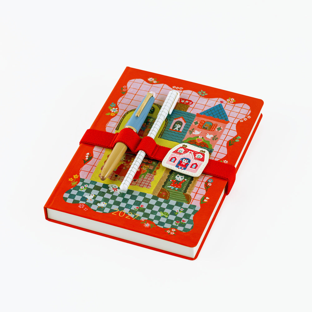 A red hardcover A5 notebook with a whimsical illustrated cover featuring a castle, checkered patterns, and a red elastic band. Two decorative pens are tucked into the cover, and a small white house-shaped charm with red illustrations is attached on the front.