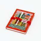 A red hardcover A5 notebook with a whimsical illustrated cover featuring a castle, checkered patterns, and a red elastic band. Two decorative pens are tucked into the cover, and a small white house-shaped charm with red illustrations is attached on the front.