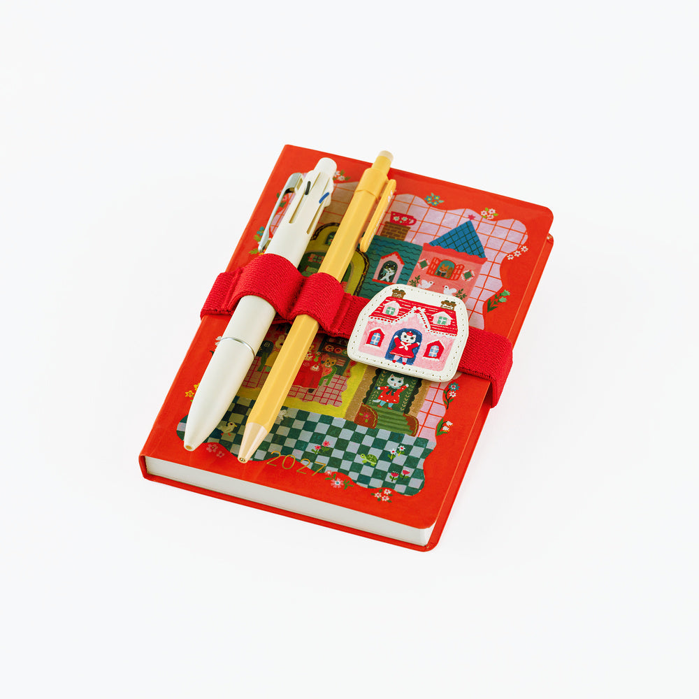 A red hardcover A6 notebook with a whimsical illustrated cover featuring a castle, checkered patterns, and a red elastic band. Two decorative pens are tucked into the cover, and a small white house-shaped charm with red illustrations is attached on the front.