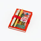 A red hardcover A6 notebook with a whimsical illustrated cover featuring a castle, checkered patterns, and a red elastic band. Two decorative pens are tucked into the cover, and a small white house-shaped charm with red illustrations is attached on the front.