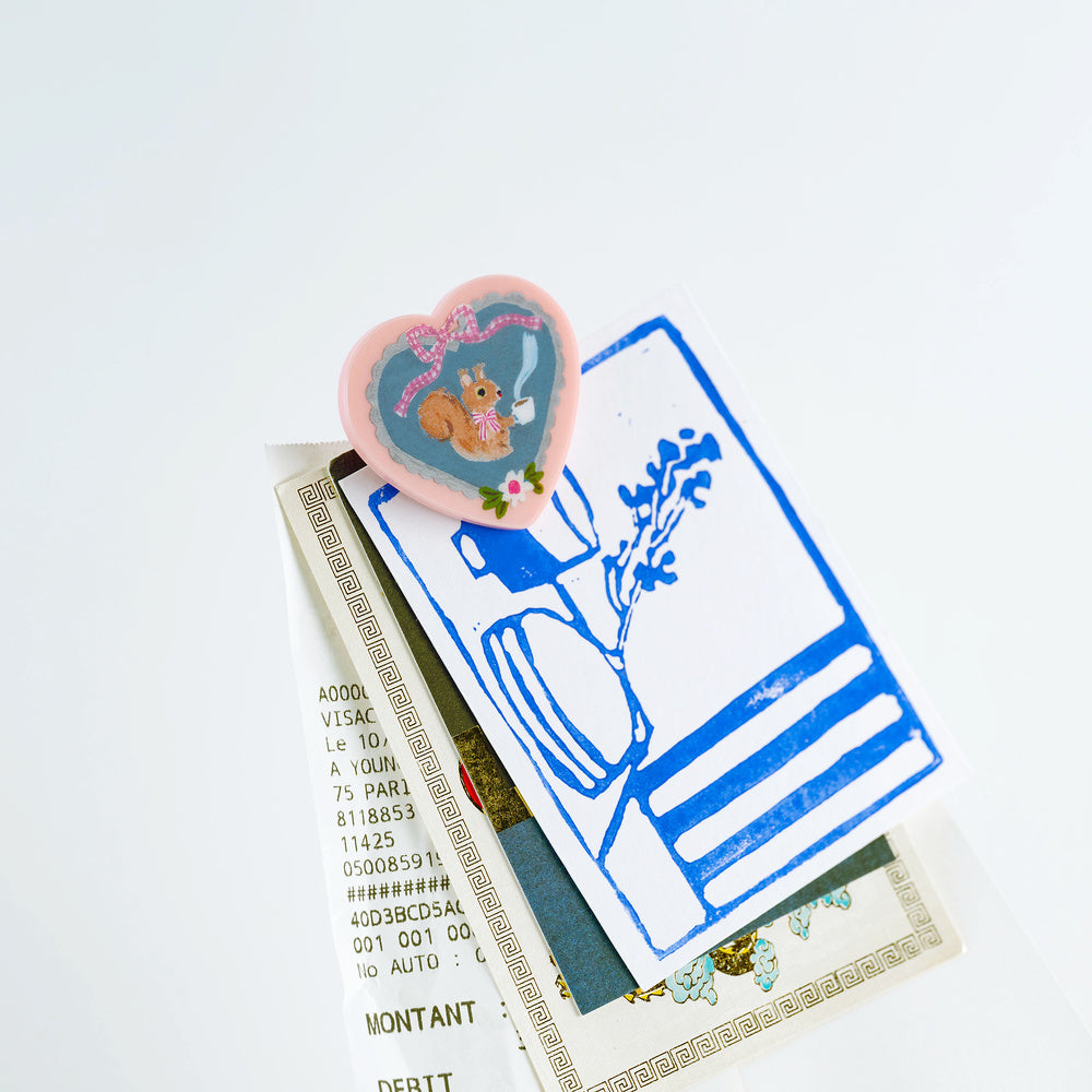 A small heart-shaped pink enamel pin featuring a cartoon squirrel holding a teacup, styled on top of vintage papers and a blue-and-white illustrated card against a clean white background.