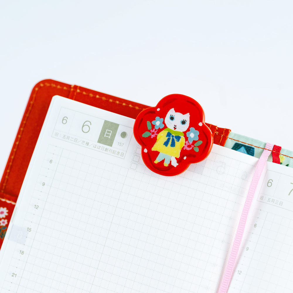 A red floral planner clip with a cartoon cat character in a yellow dress, holding an open grid planner with a pink ribbon bookmark.
